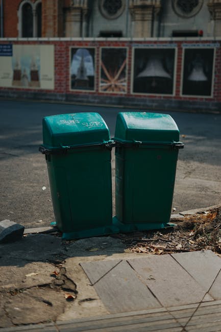 A person wearing light blue jeans and white sneakers is squatting on a paved surface, holding a broken white plastic cup with a green logo, alongside a small wooden stick. The individual's right hand